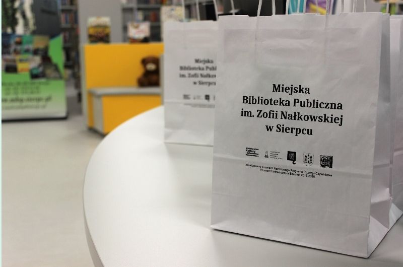 Grafika przedstawia białe papierowe torby na prezenty z napisem "Miejska Biblioteka Publiczna im. Zofii Nałkowskiej w Sierpcu" stojące na szarym blacie. W tle żółte siedziska dla dzieci oraz zielony baner Miejskiej Biblioteki.