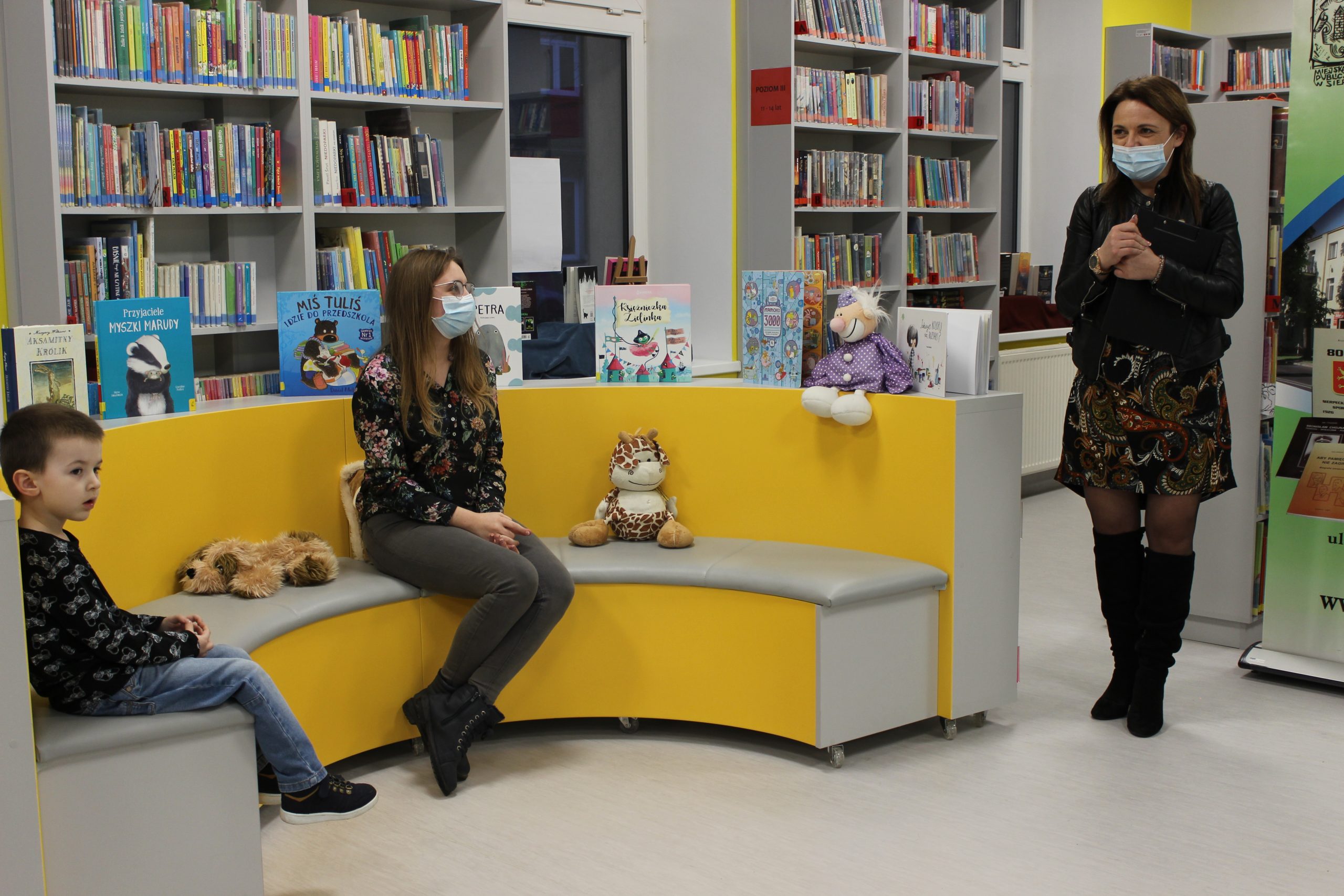 Na pierwszym planie Pani dyrektor Miejskiej Biblioteki Publicznej w Sierpcu przemawia. Na żółtych siedziskach dla dzieci siedzi młoda kobieta i mały chłopiec. W tle szare regały z książkami.