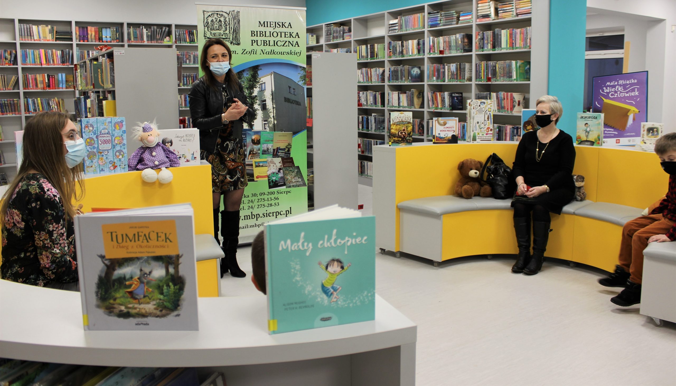 Na zdjęciu są cztery osoby Pani dyrektor Miejskiej Biblioteki Publicznej w Sierpcu Monika Stańczak oraz troje zwycięzców konkursu na najlepszego czytelnika 2021 siedzących na żółtych siedziskach dla dzieci. W tle zielony baner Miejskiej Biblioteki Publicznej w Sierpcu oraz szare regały z książkami.