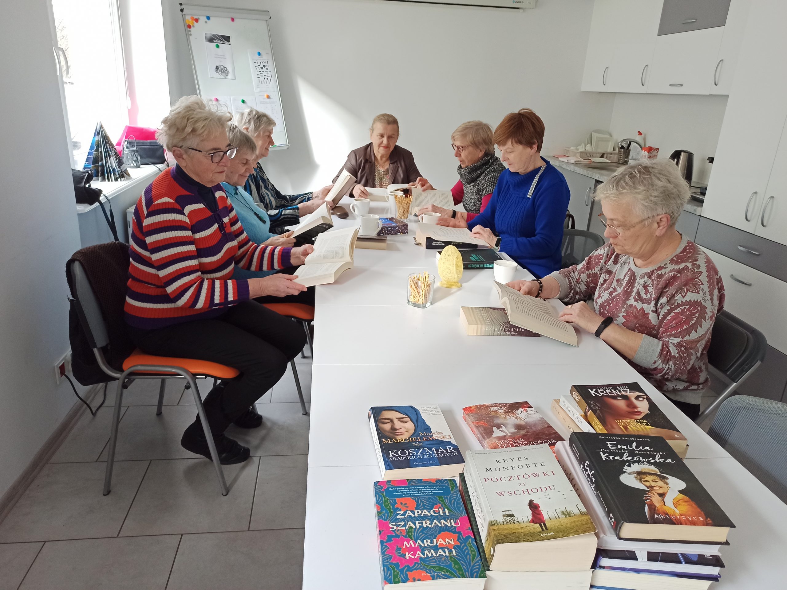 Spotkanie poświęcone literaturze w Klubie Seniora. 7 pań z Klubu czyta książki. Na pierwszym planie nowości książkowe.